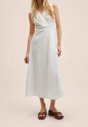 Femme portant une robe portefeuille blanche sans manches et des sandales à plateforme marron sur un fond beige uni.