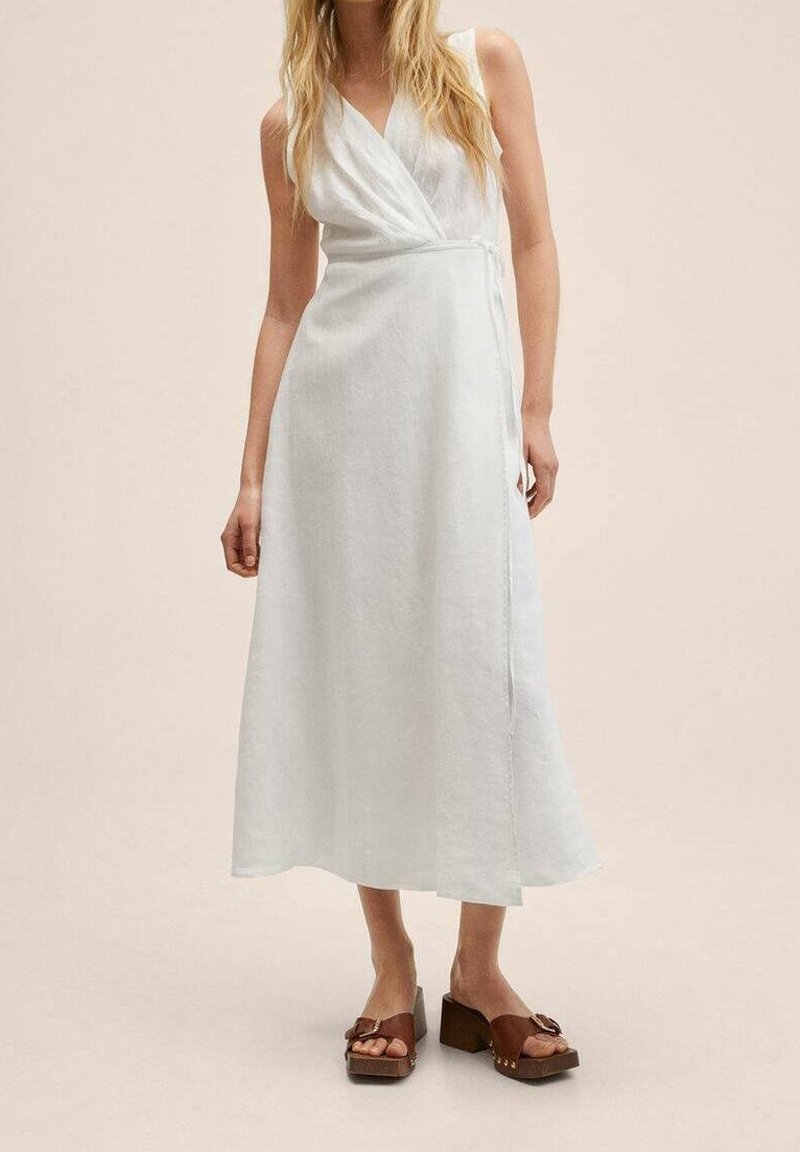 Femme portant une robe portefeuille blanche sans manches et des sandales à plateforme marron sur un fond beige uni.