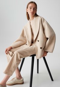 Vrouw zittend op een zwarte stoel, gekleed in een beige oversized jas, een bijpassende sweater, broek en sneakers, tegen een effen lichte achtergrond.