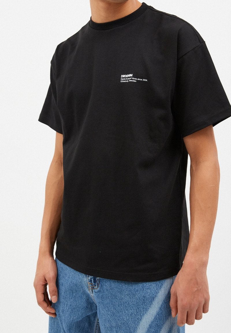 Camiseta negra de manga corta hecha de algodón, con un logo blanco en el pecho. El corte es relajado con un diseño de cuello redondo.