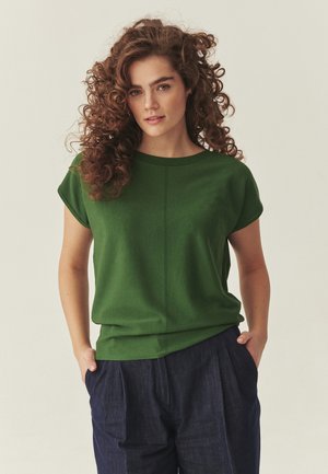 Woman with curly brown hair wearing a green short-sleeve top and dark blue pants with hands in pockets against a neutral background.