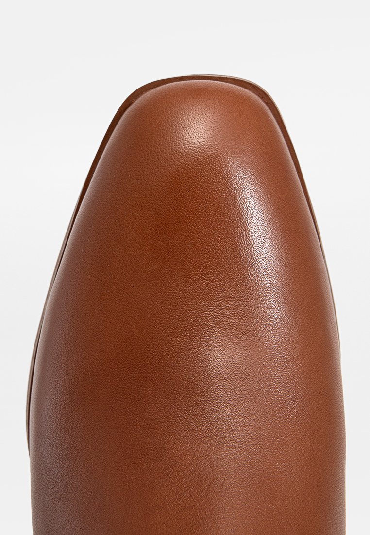 Gros plan sur la pointe d'une chaussure en cuir lisse, beige, de forme carrée arrondie avec une texture fine, sur un fond blanc uni.