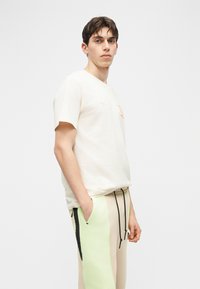 Νέος άνδρας με κοντά σκούρα μαλλιά, που φορεί λευκό κοντομάνικο μπλουζάκι και μπεζ παντελόνι με πράσινες και μαύρες πλευρικές λωρίδες, στέκεται με τα χέρια στις τσέπες.