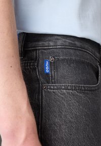 Sorte denim jeans med en blå "HUGO" label på baglommen, der har et tætsiddende design og en subtil tekstur.