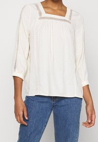Weiße Bluse mit quadratischer Ausschnitt, strukturiertem Stoff und Dreiviertelärmeln, die mit Spitzenbesatz verziert sind. Getragen mit blauen Jeans.