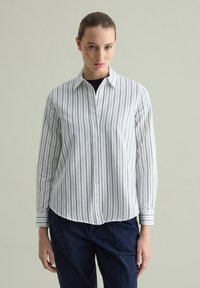 Camicia bianca con righe verticali blu navy. Presenta un colletto classico e maniche lunghe con polsini a righe. Tessuto di cotone morbido.