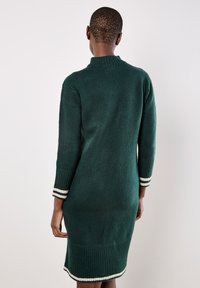Abito lavorato a maglia verde con collo alto e maniche lunghe. Presenta polsini e orlo a coste con strisce bianche. Design aderente e testurizzato.