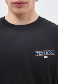 Κοντινό πλάνο ενός ατόμου που φοράει μαύρο πουκάμισο με το κείμενο "TOMMY" σε μπλε χρώμα και ένα μικρό λογότυπο Tommy Jeans με κόκκινο, λευκό και μπλε στην π εριοχή του στήθους.