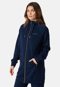 Felpa con zip colore navy realizzata in morbido materiale, dotata di cappuccio, zip dorata, tasche e la scritta "SEASIDE" ricamata sul petto.
