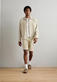 Chemise beige clair à boutons sur un t-shirt blanc. Shorts beiges et chaussures marron. Le modèle se tient sur un sol en bois avec un fond simple.