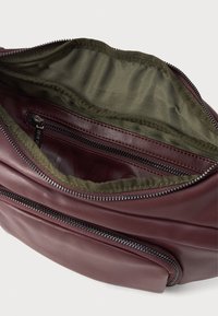 Intérieur d'un sac en cuir bordeaux avec doublure vert olive, comportant une poche zippée et un compartiment principal spacieux. Fini texturé et élégant.