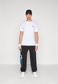 Biela tričko s krátkymi rukávmi s logom "BOSS", čierne pletené nohavice, biele tenisky a čierna čiapka. Drží tenisovú raketu s modrými a bielymi akcentmi.