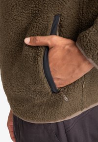 Green fleece jacket featuring a textured surface, side zip pocket with navy trim, and a gray-lined hem. Hand resting in the pocket.