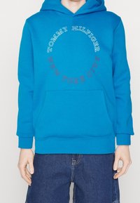 Sudadera azul con capucha y bolsillo tipo canguro, con texto circular en blanco y rojo que dice "TOMMY HILFIGER NUEVA YORK." Tejido de tela suave.