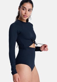 Maillot de bain noir à manches longues avec un détail noué à la taille, présentant une découpe, et une texture de tissu lisse.
