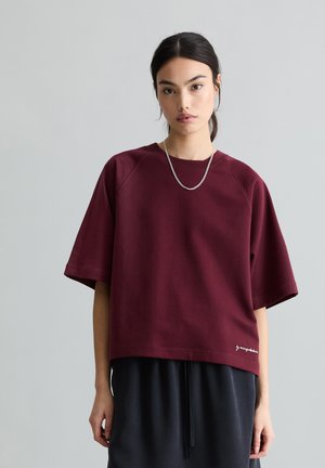 Jeune femme aux cheveux noirs attachés en queue de cheval basse, portant un haut ample à manches courtes de couleur bordeaux et un pantalon noir, sur un fond gris uni.