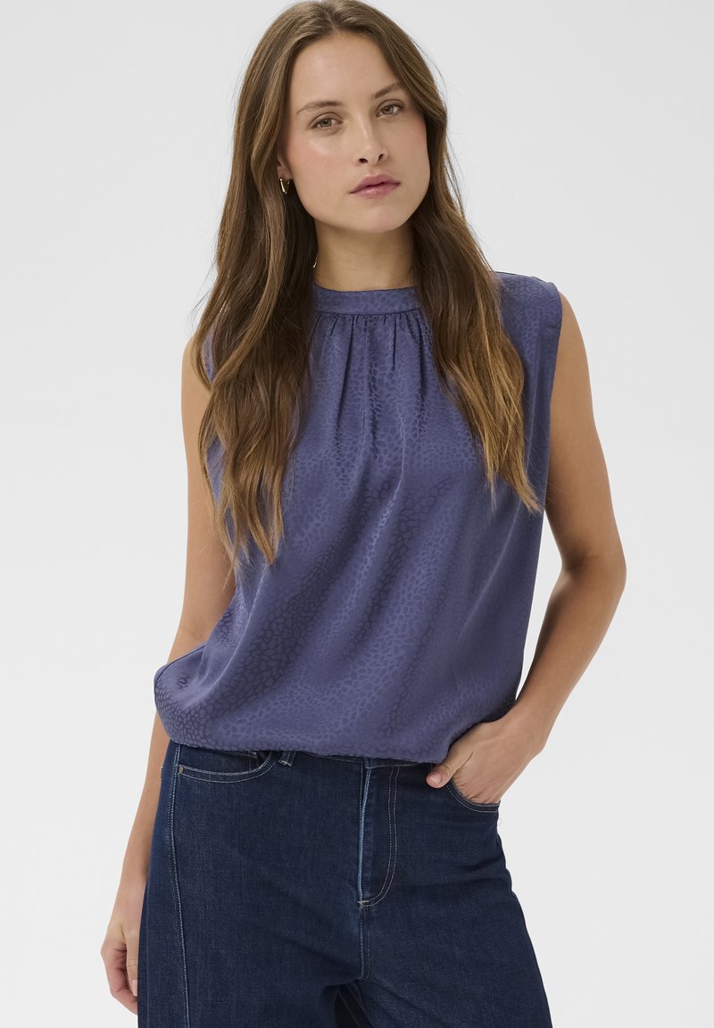 Frau mit langem braunem Haar trägt ein ärmelloses, blau gemustertes Oberteil, das in dunkelblaue Jeans gesteckt ist, und posiert mit einer Hand in der Tasche.