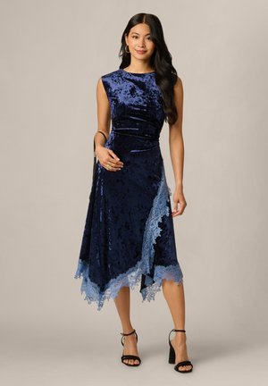 Donna con lunghi capelli scuri che indossa un abito senza maniche in velluto blu navy con bordatura in pizzo azzurro chiaro e sandali neri con tacco alto, in piedi davanti a uno sfondo semplice.