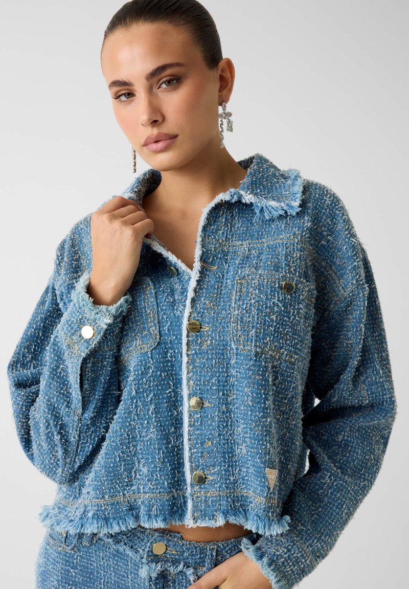 Blauwe geaderde denimjas met rafelige randen en twee voorzakken, voorzien van goudkleurige knopen en een kraagontwerp.