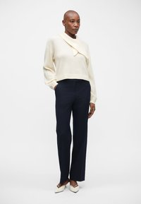 Sudadera recortada de marfil con un cuello drapeado, combinada con unos pantalones rectos azul marino y unas bailarinas blancas de punta. Textura suave, diseño minimalista.