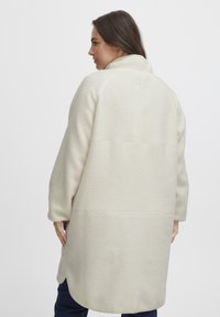 Krämfärgad fleecejacka med hög krage, långa ärmar och en texturerad yta. Har en avslappnad passform och en rundad nederkant.