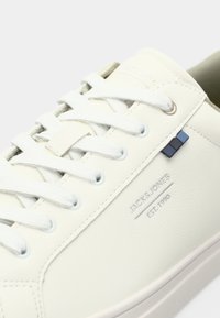 White leather sneaker with white laces, subtle stitching, and "JACK & JONES EST. 1990" printed on the side near a small blue tag.