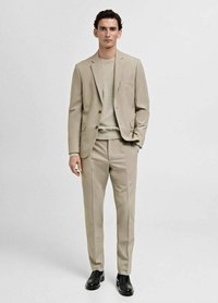 Beige kostym med en skräddarsydd jacka och slim fit byxor, i kombination med en ljus tröja. Har en slät yta och eleganta svarta skor.