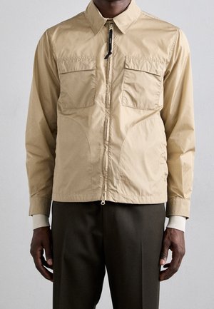 Homme portant une veste beige zippée avec deux poches à rabat sur le devant, superposée à un pull blanc à poignets côtelés, associée à un pantalon foncé.