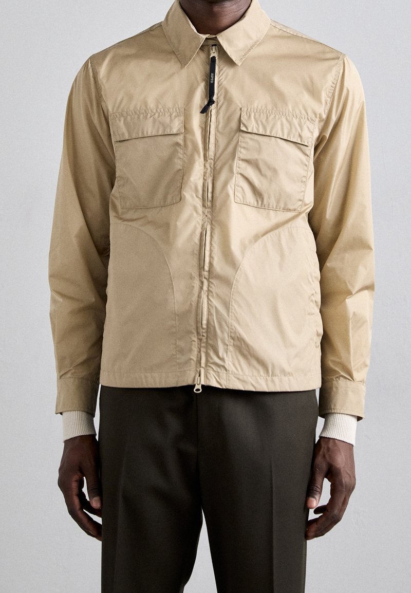 Homme portant une veste beige zippée avec deux poches à rabat sur le devant, superposée à un pull blanc à poignets côtelés, associée à un pantalon foncé.