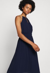 Rochie midi pliată de culoare bleumarin, cu guler halter și talie elastică, având un detaliu cu decupaj în față și o formă fluidă.