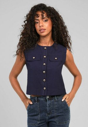 Femme aux cheveux bouclés portant un haut bleu marine sans manches boutonné et un jean taille haute bleu foncé, les mains dans les poches, debout devant un fond gris.