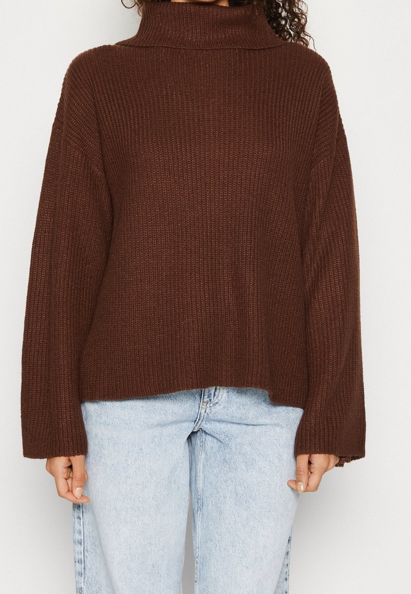 Suéter de punto marrón con cuello alto, de corte holgado y textura acanalada, combinado con pantalones vaqueros de pierna ancha en azul claro.