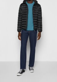 Sort puffersjakke med hætte, blå T-shirt, mørke jeans og sorte sneakers. Jakken har vandret quilting og lynlås lukning.