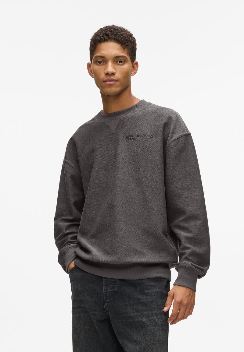 Sivá mikina s okrúhlym výstrihom, textúrovaná látka, rebrované manžety a malé logo na hrudi, v kombinácii s tmavými džínsami.