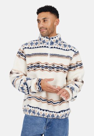 Uomo che indossa un pullover in pile bianco con motivi geometrici blu e marroni, colletto con zip a metà, e jeans blu, che guarda di lato.