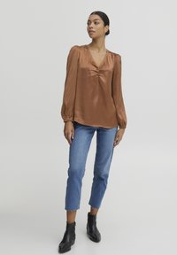 Blusa in satin marrone con scollo a V e dettagli arricciati, abbinata a jeans cropped blu chiaro e stivaletti neri alla caviglia con finitura testurizzata.