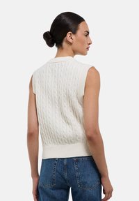 Pull en maille torsadée sans manches de couleur blanc cassé avec un motif texturé. Ourlet et col côtelés ; mannequin portant avec un jean bleu foncé.