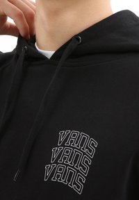 Svart hoodie med staplad vit "VANS" logotypdesign. Dra åt huva och mjuk textur. Närbild av halsringning och logodetaljer.