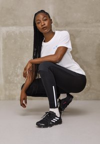 Maglietta atletica bianca a maniche corte con logo blu, leggings neri aderenti con strisce bianche ai lati e scarpe sportive nere con dettagli bianchi.