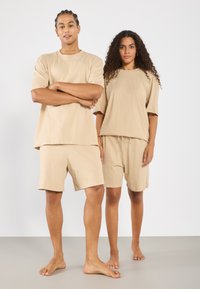 Beige pamut pólók rövid ujjakkal, azonos színű rövidnadrággal párosítva. Laza szabás, minimalista design és puha textúra, mezítláb viselve egy világos háttér előtt.
