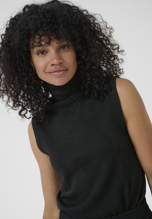 Frau mit lockigem schwarzen Haar, die einen ärmellosen schwarzen Rollkragenpullover vor einem schlichten hellen Hintergrund trägt.
