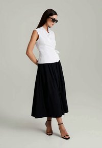 White sleeveless top with a textured finish, cinched waist, and ruffled collar paired with a black, flowy, ankle-length skirt. Black heels.