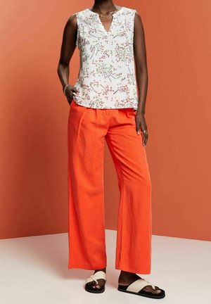 Ærmeløs top med blomsterprint i hvid, kombineret med brede bukser i lys orange. Model iført sorte sandaler med hvide remme.