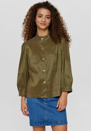 Olijfgroene satijnen blouse met een hoge kraag, knoopsluiting en pofmouwen; gecombineerd met een spijkerrok. Bevat twee voorzakken.