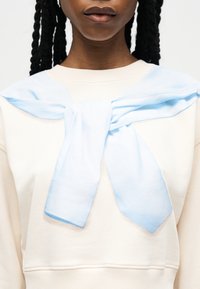 Κοντινό πλάνο ενός ατόμου που φοράει μια εκρού φούτερ μπλούζα με ένα ανοιχτό μπλε φούτερ δεμένο γύρω από τους ώμους του σε φιόγκο.