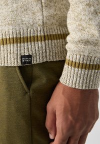 Beige strikkegenser med grønne striper ved kanten og ermene, sammen med mørk olivengrønne bukser med sidelomme.