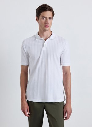 Giovane uomo con capelli corti castani che indossa una polo bianca e pantaloni verde oliva, in piedi davanti a uno sfondo chiaro e uniforme.