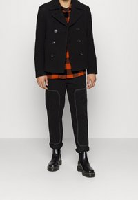 Veste noire croisée en tissu texturé portée sur une chemise à carreaux orange et noir. Pantalon noir avec coutures blanches et bottes noires.