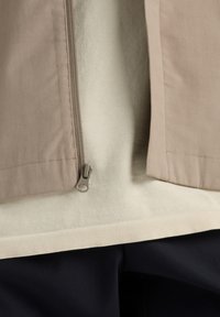 Beige jack met een rits, gelaagd over een lichtgekleurde trui. Donkere, gemêleerde broek zichtbaar eronder.