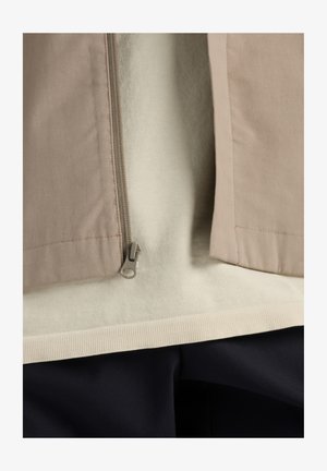 Šviesiai rudas švarkas su užtrauktuku, dėvimas ant šviesaus megztinio. Apačioje matosi tamsios, tekstūruotos kelnes.